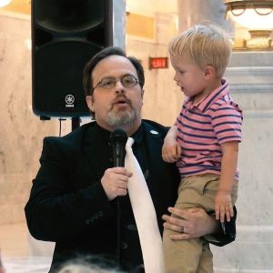 Paul Gibbs speaks into microphone while holding son