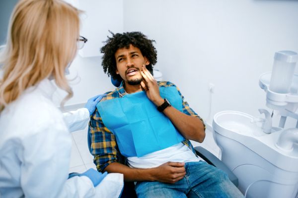 Man in a Dental Chair Touches His Jaw While a Clinician Listens Nearby, During a Checkup. (informational)