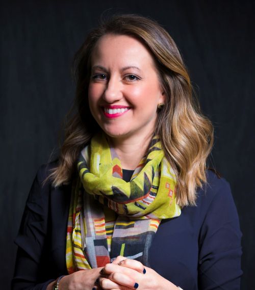 Julie Seward Headshot – CareQuest Institute Portrait of a Smiling Woman with Blonde Highlights, Wearing a Navy Blouse and a Yellow Patterned Scarf Against a Dark Backdrop.