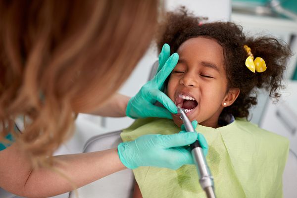 Girl at a Dental Checkup with a Dentist Using a Dental Tool While the Patient Opens Her Mouth; Both Wearing Gloves and a Green Bib.