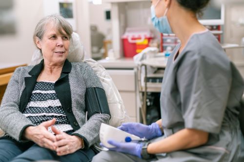 Older Woman Sits in a Hospital Bed with a Blood Pressure Cuff on Her Arm, Talking with a Nurse Wearing a Mask and Gloves.