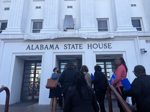People Entering the Alabama State House Entrance, a White Neoclassical Building with Columns and the Words 'alabama State House' Above the Doors, Several Individuals Carrying Blue Folders and Bags.
