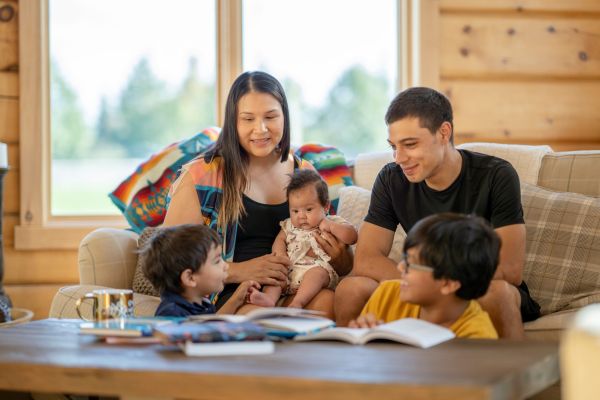 Family of Five on a Sofa in a Bright Wooden Living Room, Parents with Three Children Gathered Around an Open Book on the Coffee Table, Baby in Mom's Arms.