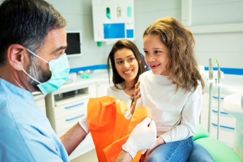 Dentist Wearing a Mask and Gloves Hands an Orange Gift Bag to a Smiling Girl in a Bright Dental Clinic, As an Assistant Watches.