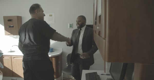 Two Men Shake Hands in a Small Office: One in Black Scrubs and the Other in a Suit, Near a Desk with a Computer and Cabinets Nearby.