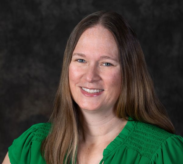 Smiling Woman with Long Brown Hair in a Green Blouse Against a Dark Textured Background, Posing for a Portrait.