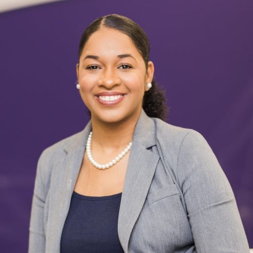 Smiling Professional Portrait of a Woman in a Gray Blazer and Pearl Necklace Against a Purple Backdrop.
