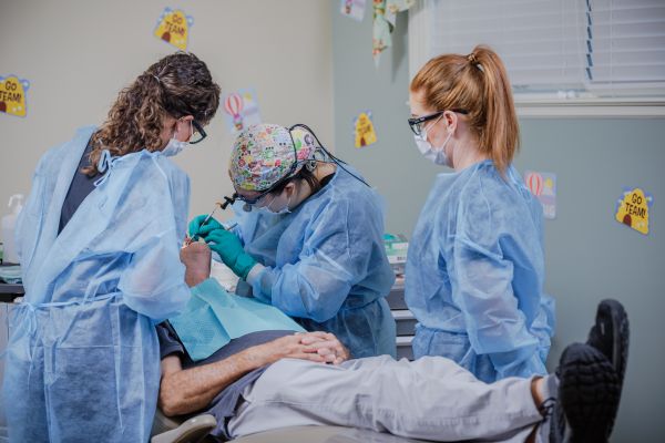 Access Dental exam – CareQuest Institute Three Clinicians in Ppe Perform a Dental Procedure on a Patient Reclining in a Chair in a Clinic.