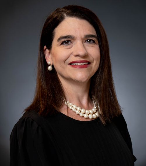 Betsy White Access Dental – CareQuest Institute Professional Headshot of a Smiling Woman with Dark Hair, Wearing a Black Top and Pearl Necklace Against a Gray Backdrop.