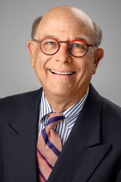 Bill Milner Headshot – CareQuest Institute Professional Headshot of an Older Man in a Navy Suit, Striped Shirt, and Pink Striped Tie, Smiling Against a Gray Background.