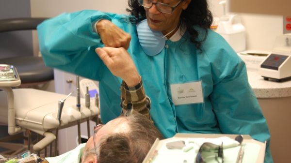 Healthcare Professional in Turquoise Protective Gown and Mask Lifts a Patient’s Head During an Exam in a Treatment Room.