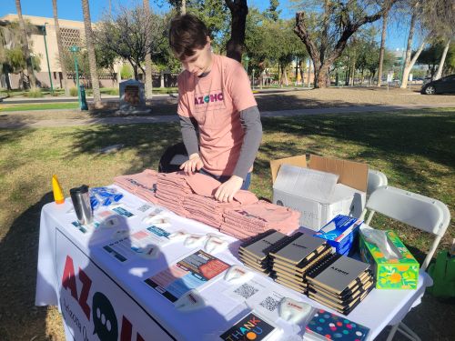 Young Man in a Pink Shirt Organizes Books and Flyers on an Outdoor Table in a Sunny Park, with a Banner and Chairs Nearby.