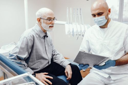 Older Man in a Dental Chair Listens As a Clinician in a Mask and Gloves Shows Him a Dental X-ray Image.