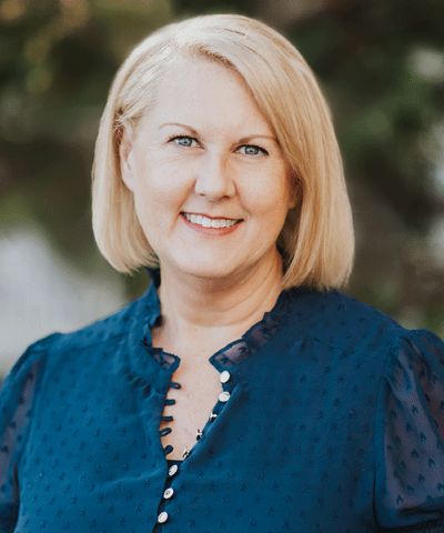 Portrait of a Smiling Blonde Woman in a Blue Blouse Outdoors with a Blurred Green Background.