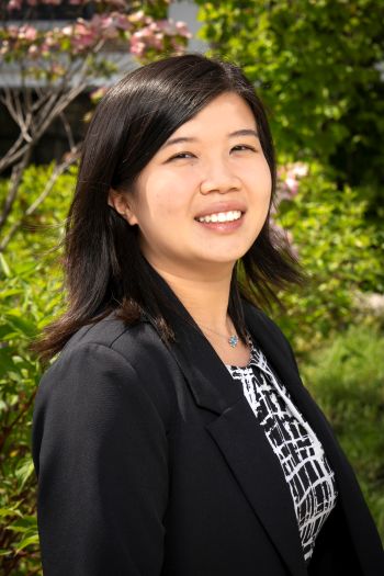 Smiling Woman Outdoors, Wearing a Black Blazer and Patterned Blouse, with Greenery in the Background.