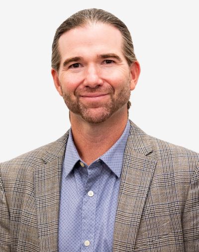 Smiling Man with Shoulder-length Gray-brown Hair and a Trimmed Beard, Wearing a Light Blue Button-down Shirt and Brown Plaid Blazer, Posed for a Professional Head-and-shoulders Portrait Against a Light Background.