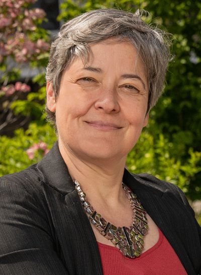 Andrea Clark Headshot – CareQuest Institute Smiling Woman with Short Gray Hair in a Black Blazer and Red Top, Wearing a Chunky Necklace, Outdoors with Greenery Behind Her
