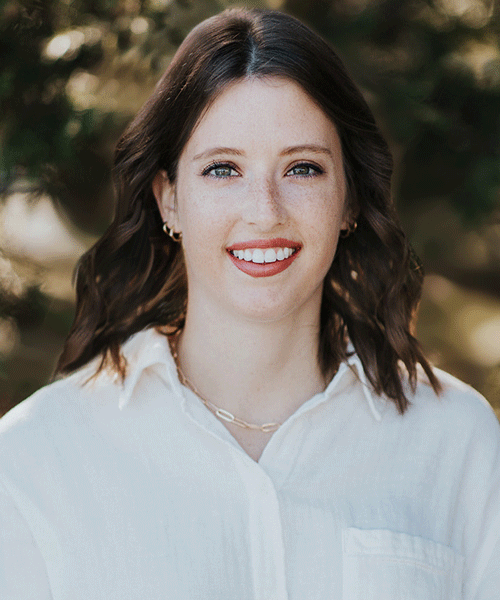 Caroline-McLeod-Web – CareQuest Institute Portrait of a Smiling Woman with Shoulder-length Dark Hair in a White Button-down Shirt Outdoors.