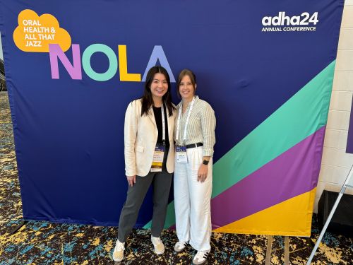 Two Women Posing in Front of a Colorful Banner Reading Nola at the Adha24 Annual Conference.
