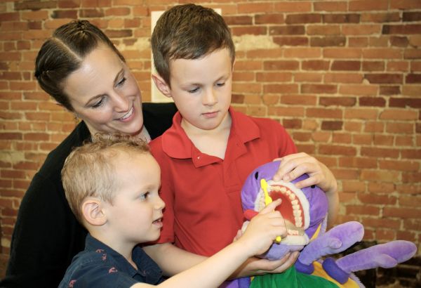Mother and Two Young Boys Examine a Purple Plush Toy with an Open Mouth, As One Child Points to Its Teeth During a Hands-on Demo.