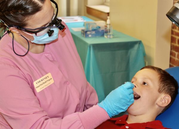 Dental Hygienist in Pink Scrubs, Mask, and Gloves Examining a Smiling Boy's Mouth During a Dental Check-up in a Clinic.