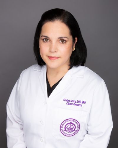 Head-and-shoulders Portrait of a Female Clinician in a White Lab Coat with a Purple Circular Emblem on the Pocket, Standing Against a Gray Background, Conveying a Professional Headshot.