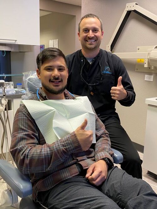 Patient in a Dental Chair Giving a Thumbs Up, with a Clinician Standing Behind Him in a Dental Clinic.
