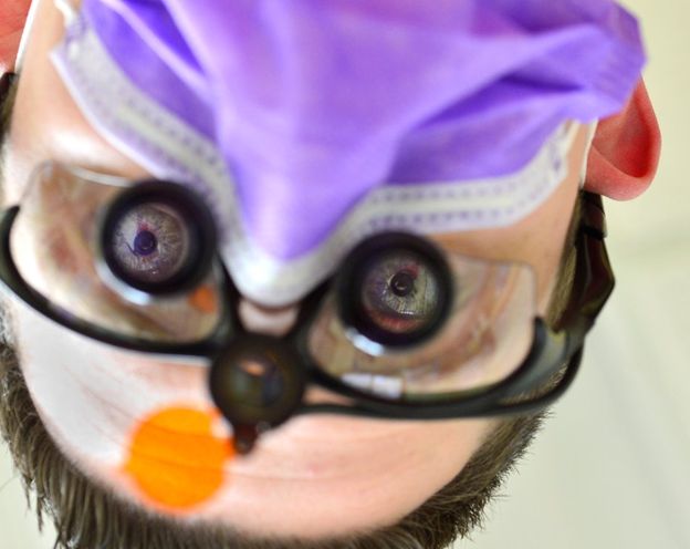 Close up of glasses – CareQuest Institute Close-up of a Medical Professional Wearing a Purple Surgical Cap, Mask, and Loupe Glasses, with an Orange Prop Nose Visible, Looking Toward the Camera (photo Upside Down).