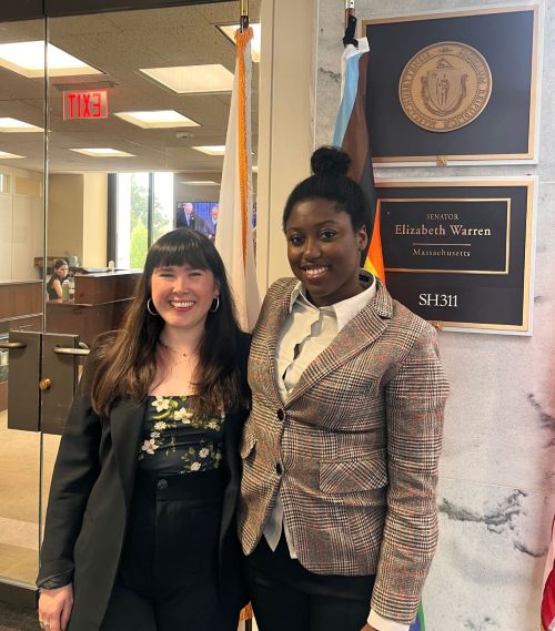 Two Women Smiling for a Photo Inside a Government Office, with a Plaque for Senator Elizabeth Warren and Flags Behind Them.