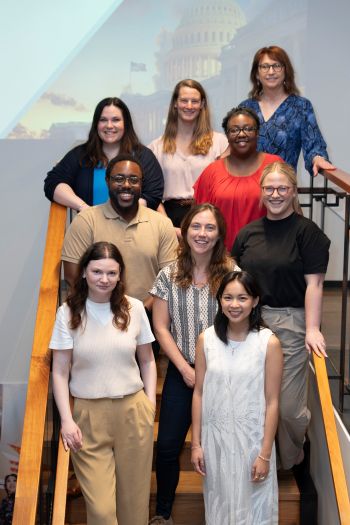 Diverse Group of Nine Adults Posing on a Wooden Staircase Indoors, Smiling at the Camera.