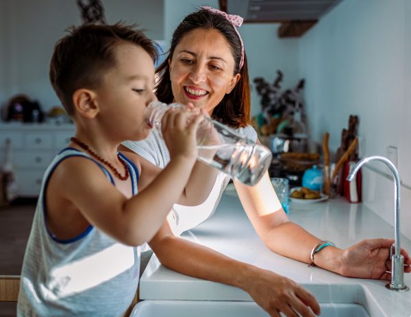 Mother Smiling As Her Young Son Drinks Water from a Bottle at the Kitchen Sink, with His Mother Nearby.