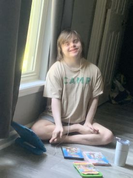 Joshua Neely Sitting – CareQuest Institute Young Person Sitting Cross-legged on a Wooden Floor by a Window, Wearing a Beige T-shirt and Earbuds, Smiling, with Children’s Books and a Mug on the Floor Nearby.