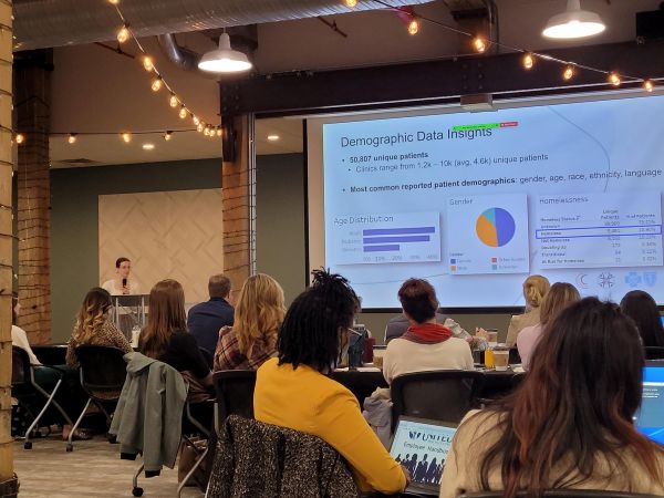 COrHT_NC_Session – CareQuest Institute Speaker at a Podium Presenting Demographic Data Slides to an Audience in a Conference Room with String Lights Overhead.