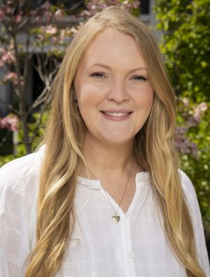 Christine Kanan Headshot – CareQuest Institute a Woman with Long Blonde Hair Wearing a White Blouse Smiles at the Camera in an Outdoor Garden Setting.
