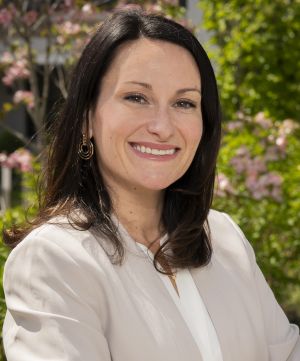 Danielle Apostolon Headshot – CareQuest Institute Professional Headshot: Smiling Woman with Dark Hair in a White Blazer, Outdoors with Greenery Behind Her.