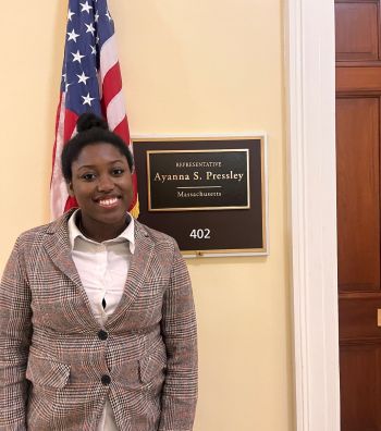 Woman in a Plaid Blazer Posing in a Hallway Beside an American Flag and a Plaque Behind Her.
