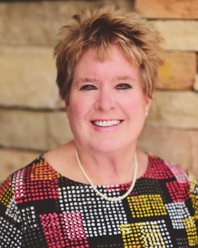 Smiling Woman with Short Blond Hair Wearing a Colorful Patterned Top and a Pearl Necklace, Standing in Front of a Brick Wall.