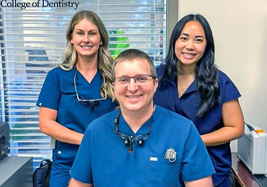 Three Dental Staff in Blue Scrubs Pose in a Clinic; Man Seated Front with Loupes, Two Women Standing Behind.