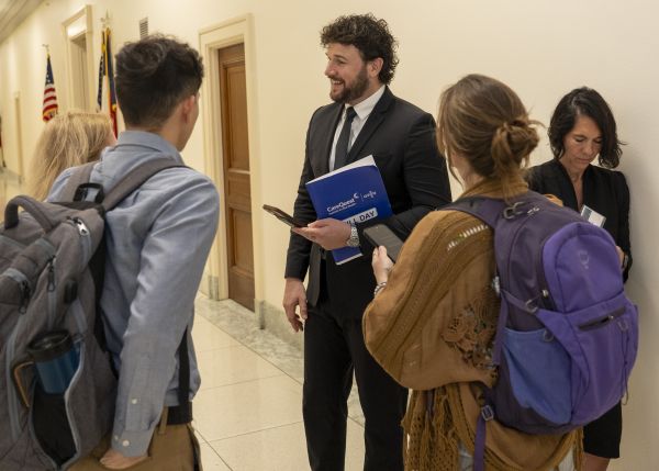 Matt Steele at OPEN Hill Day – CareQuest Institute a Man in a Suit Chats with Students in a Hallway, Holding a Blue Folder and Phone As They Gather Around Him.