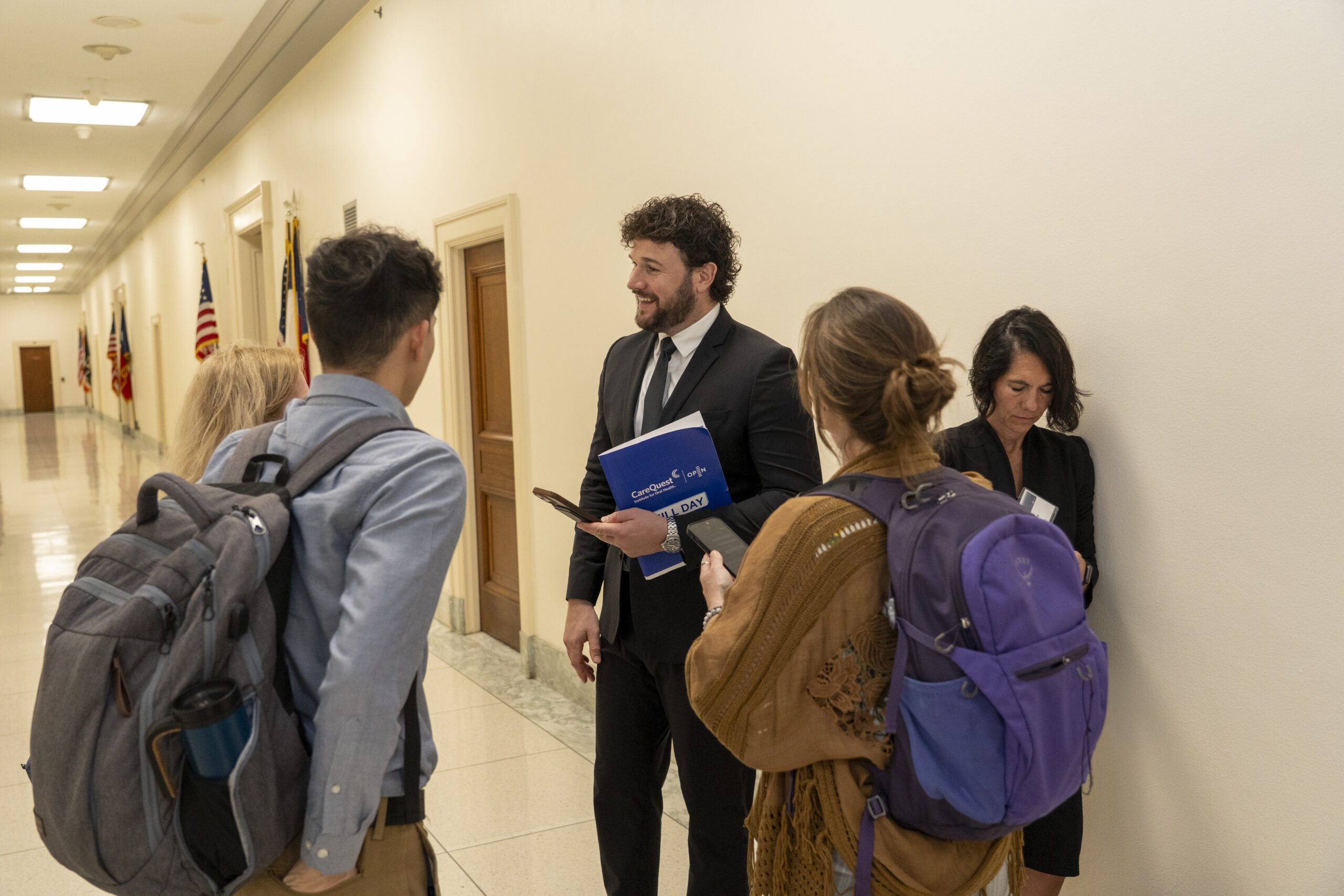 Group of Professionals Chatting in a Hallway; a Man in a Suit Hands a Blue Folder to Colleagues with Backpacks Nearby.
