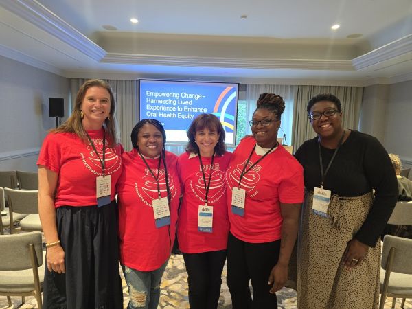 Five Women in Red Conference Shirts with Lanyards Pose Together in a Room; a Slide About Empowering Change is Visible on a Screen Behind Them.