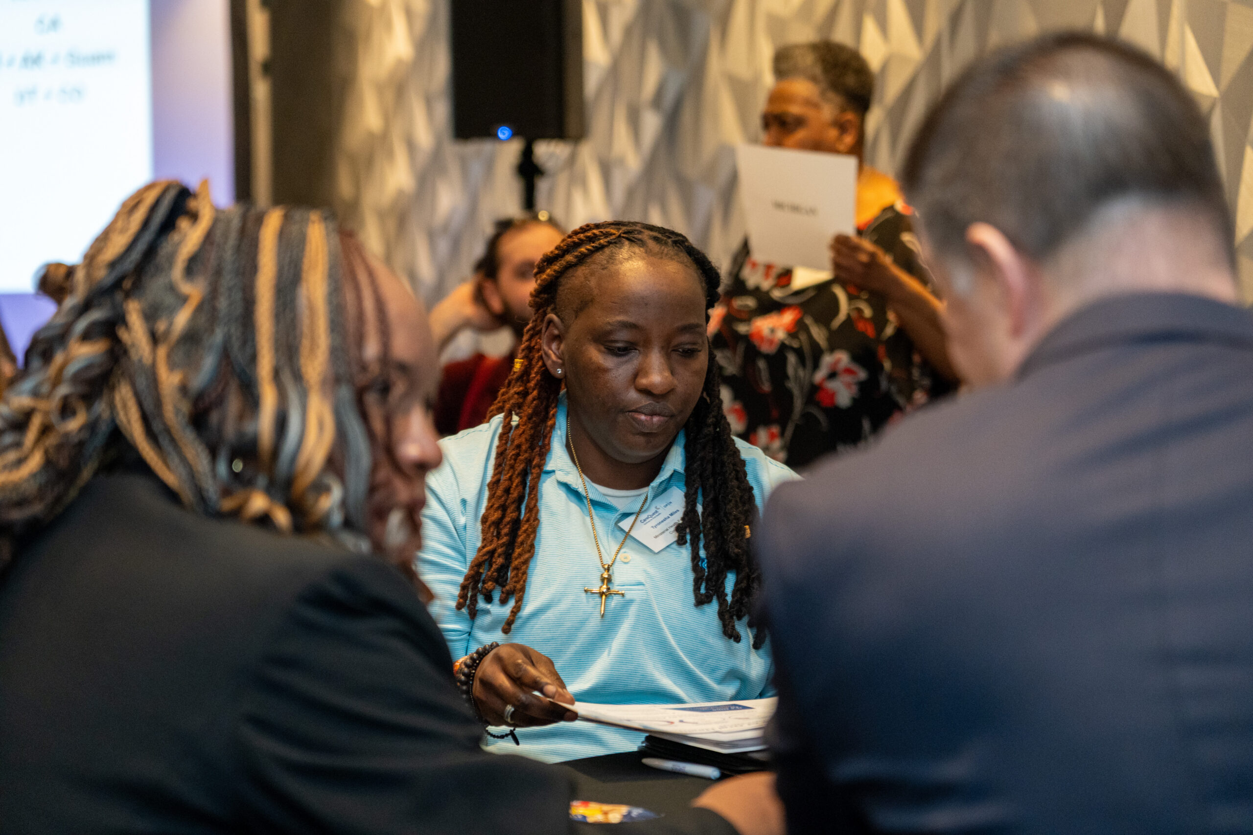 Woman with Braided Hair in a Blue Polo Reads Papers at a Busy Meeting.