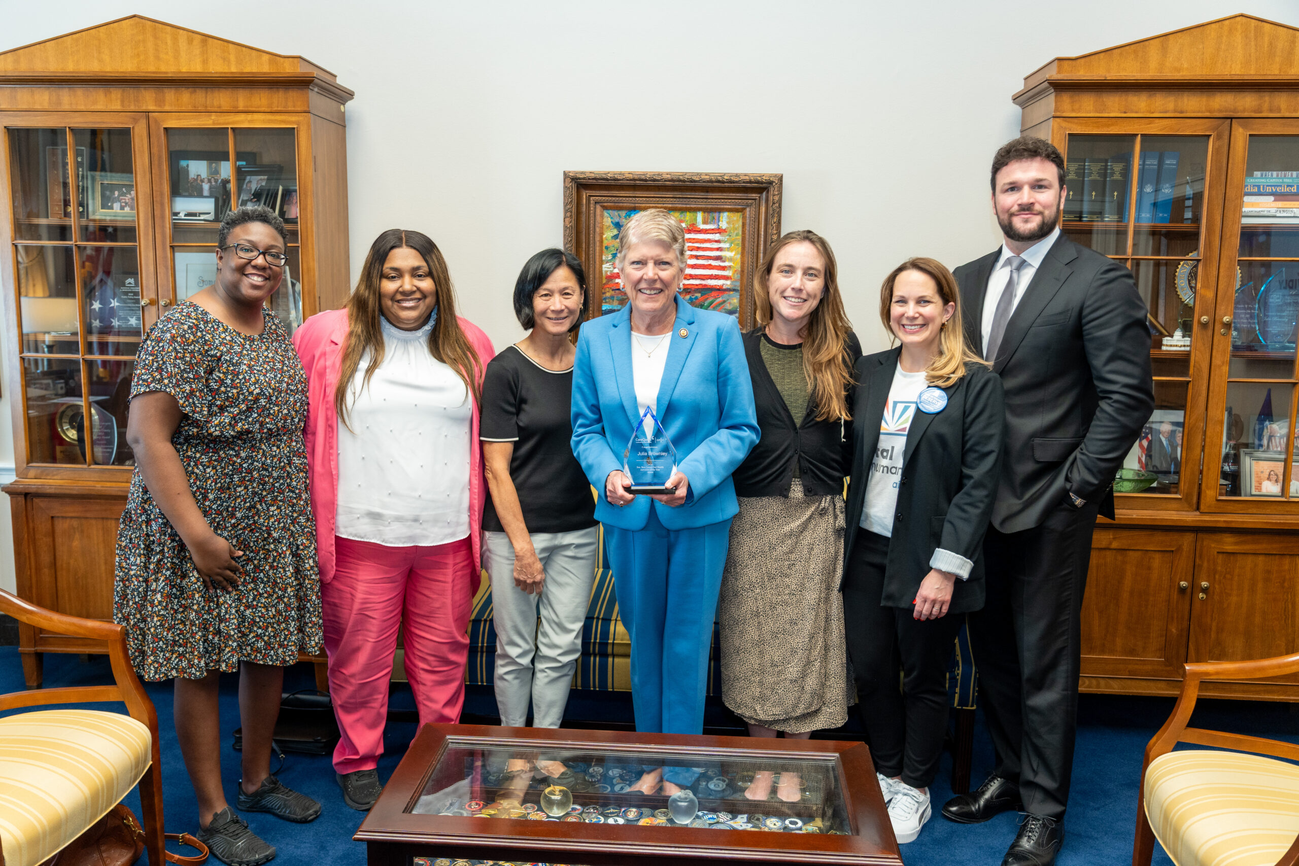 Seven Professionals Pose for a Photo in an Office, with a Framed Painting and Wood Cabinets in the Background; the Center Woman in Blue Holds a Glass Award.