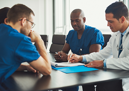 Three Healthcare Professionals—two in Blue Scrubs and One in a White Coat—discussing a Document on a Blue Folder at a Conference Table.