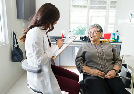 Caregiver or Clinician Sits Beside an Elderly Woman in a Bright Clinic, Writing on a Clipboard and Taking Notes.