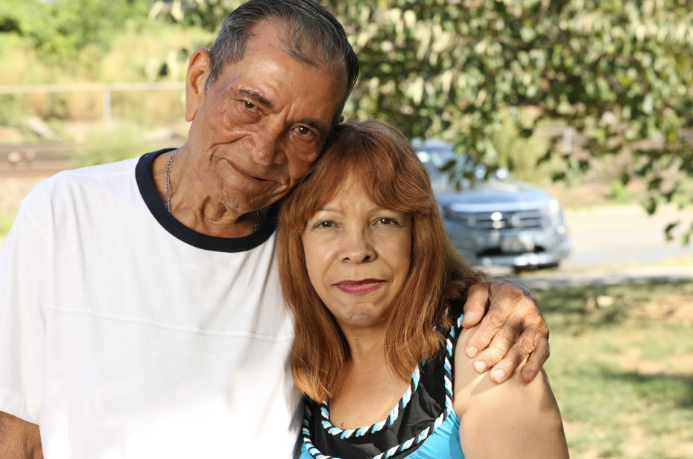 Share-Story Older Couple with Arms Around Each Other, Smiling Outdoors in a Park-like Setting with Trees in the Background.