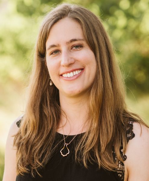 Close-up Portrait of a Smiling Woman with Long Brown Hair, Wearing a Black Top, Standing Outdoors in Soft, Sunny Light.