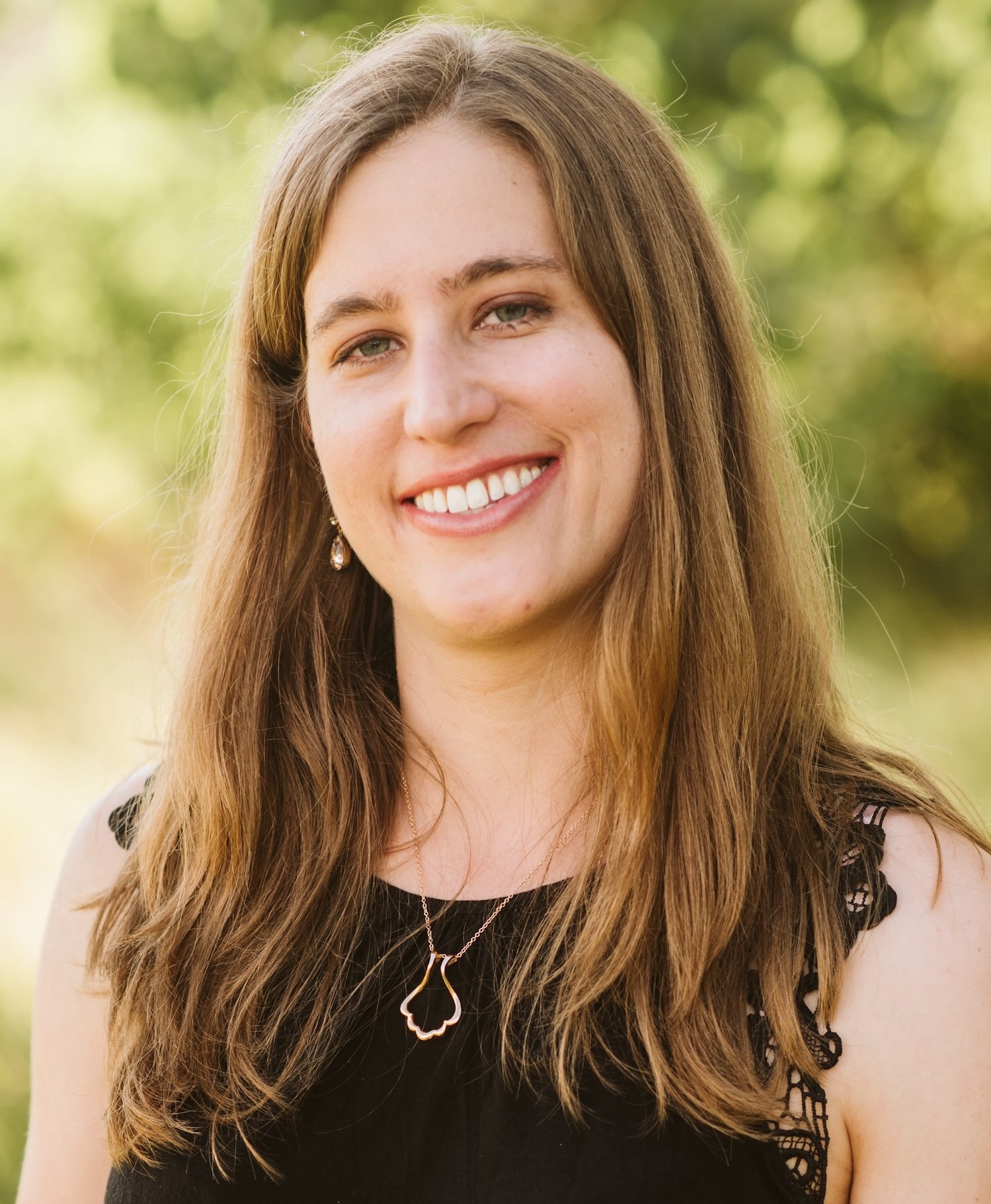 Close-up Portrait of a Smiling Woman with Long Light Brown Hair, Outdoors with a Green Blurred Background.
