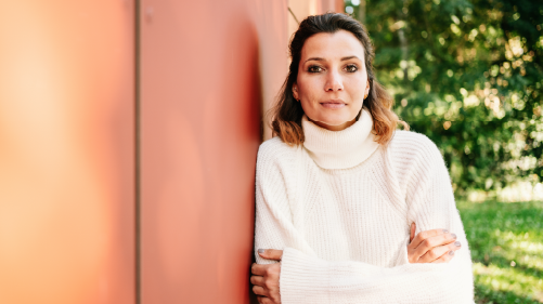 Woman with Brown Hair in a White Turtleneck Sweater Leaning Against an Orange Wall Outdoors, Looking at the Camera.