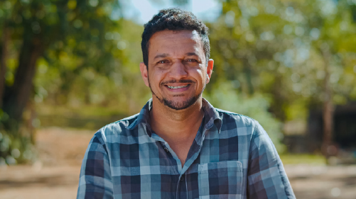 Smiling Man in a Blue Plaid Shirt Stands Outdoors in a Sunlit Park.